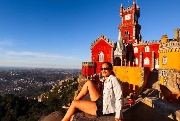 Woman wearing sunglasses sitting by colorful Pena Palace with scenic mountain view