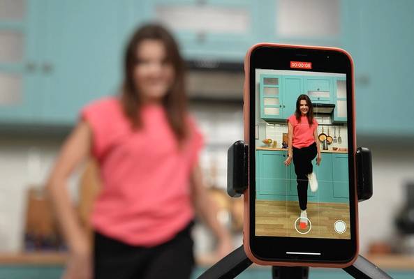 Young woman recording fitness video at home kitchen using smartphone on tripod.