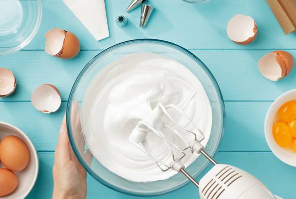 Hand mixing egg whites to stiff peaks with electric beater in a glass bowl on blue surface