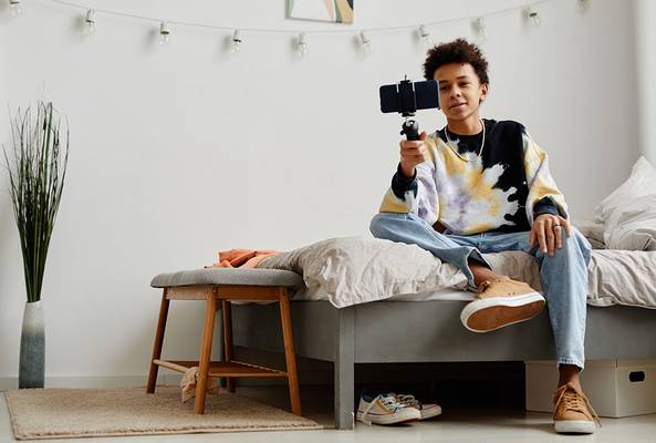 Teen sitting on bed holding smartphone on selfie stick in modern bedroom interior