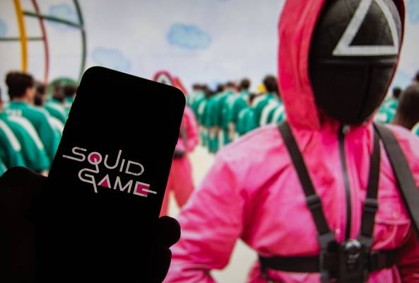 Smartphone displaying Squid Game logo with character in pink suit and green tracksuits background