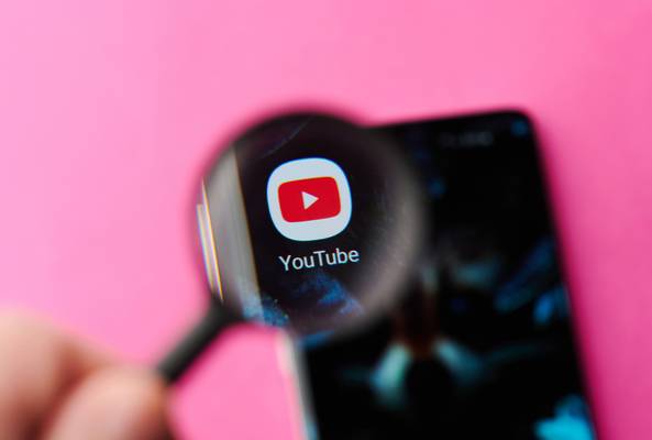 YouTube app icon magnified on smartphone screen with pink background and hand holding lens