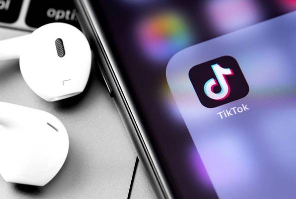 Close-up of smartphone showing TikTok app icon next to white wireless earbuds on table.
