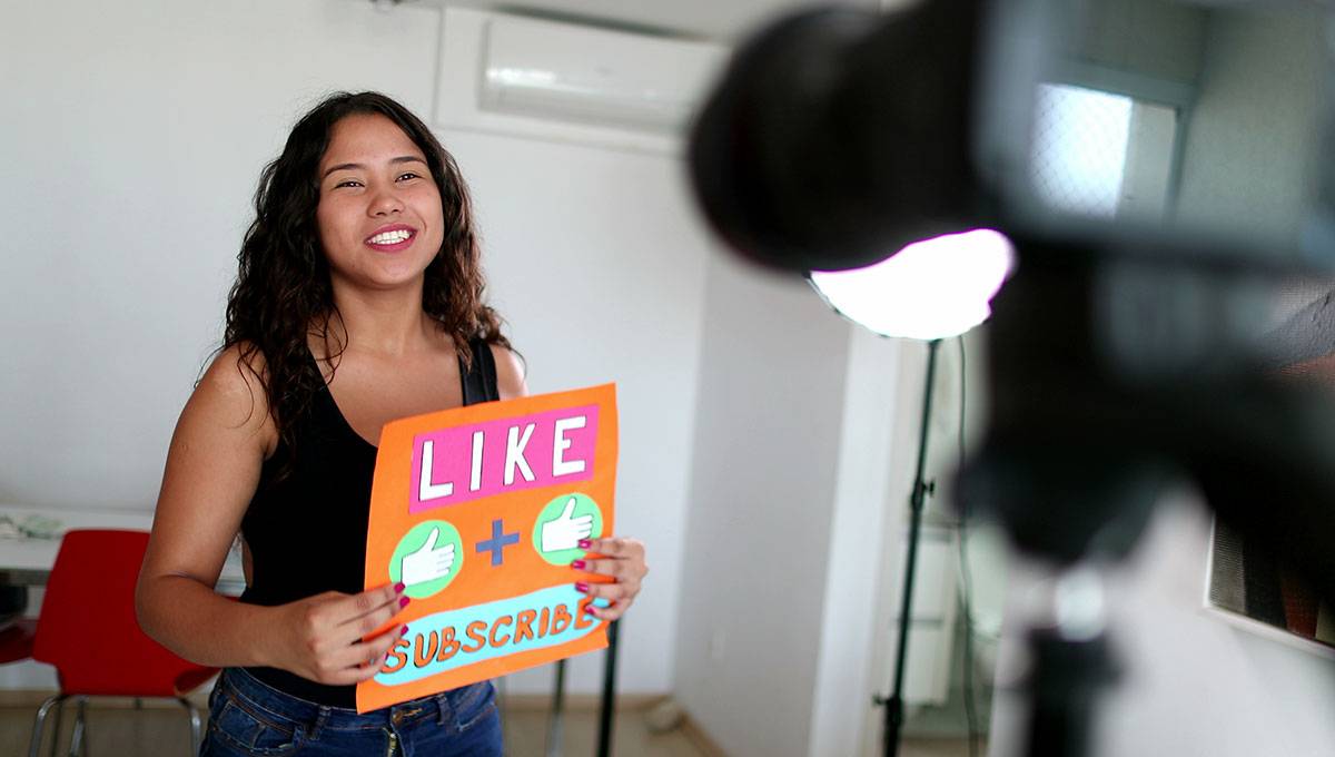 Woman holding up sign that says "like and subscribe" while filming