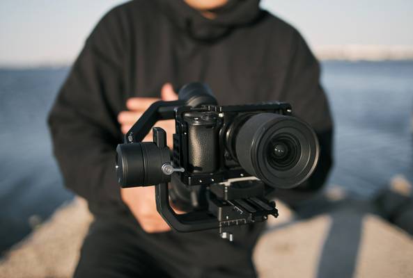 Person holding professional camera stabilizer with lens near waterfront outdoors
