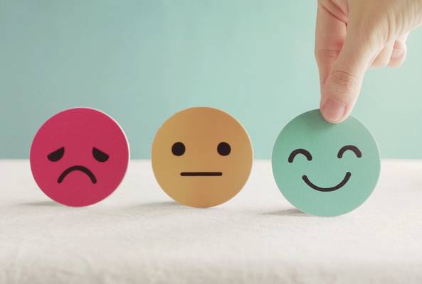 Hand picking green happy face emoji among sad and neutral emoticons on table surface.