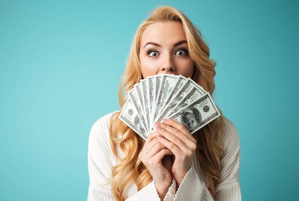 Woman holding fan of US 100 dollar bills with surprised expression on blue background