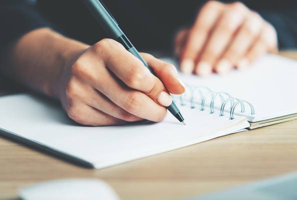 Close-up of hand writing with pen on blank spiral notebook on wooden desk