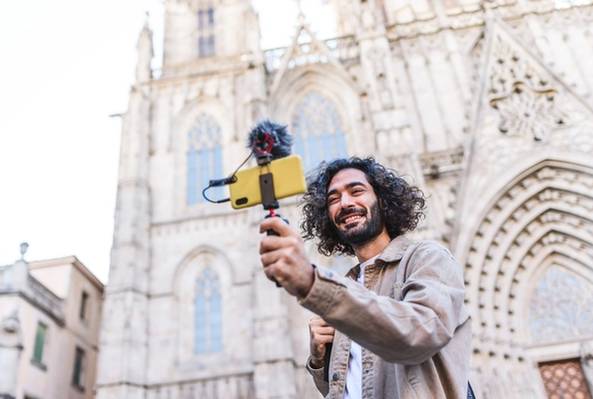 Smiling man recording travel vlog with smartphone and microphone in front of cathedral facade