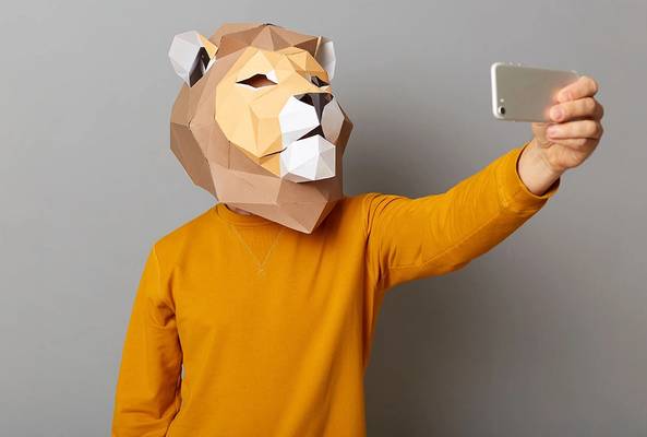 Person in orange sweater wearing a geometric lion mask taking a selfie with smartphone
