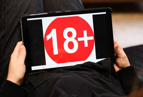 Person holding a tablet displaying a red 18+ age restriction warning symbol on screen.