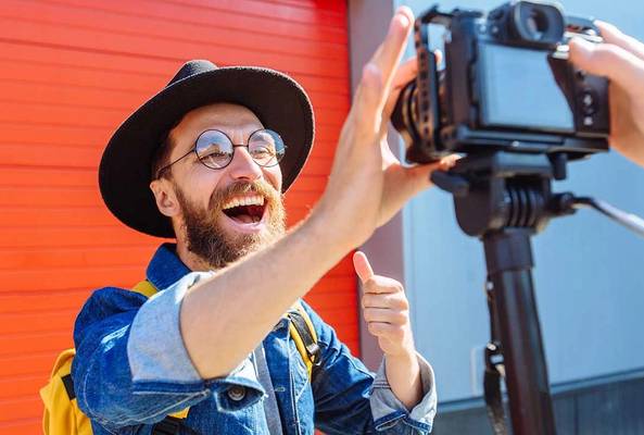Man in hat and glasses recording vlog with camera against red wall background
