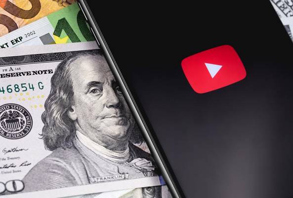Smartphone with YouTube logo on screen laying on various international banknotes and dollars.