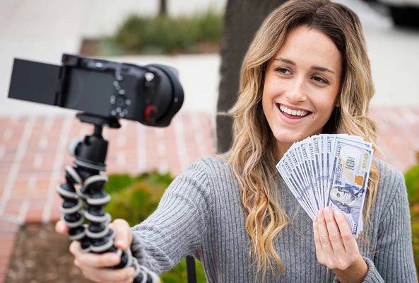 Woman filming vlog holding fan of hundred-dollar bills outdoors with camera on tripod
