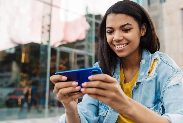 Young woman smiling while playing mobile games on blue smartphone outdoors