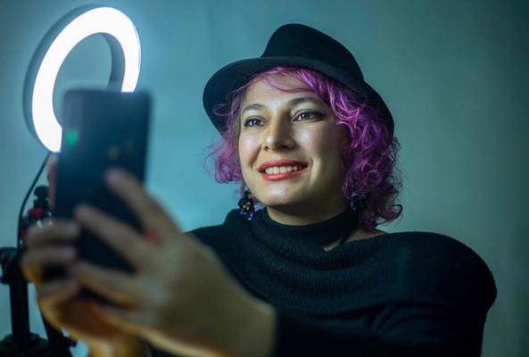 Woman with purple hair and black hat taking a selfie using a ring light for better lighting