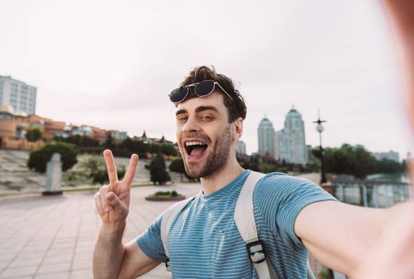 Young man smiling and making peace sign while taking selfie outdoors in cityscape