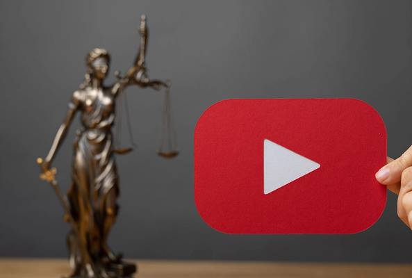 Hand holding red YouTube play button cutout with blurred Lady Justice statue in background