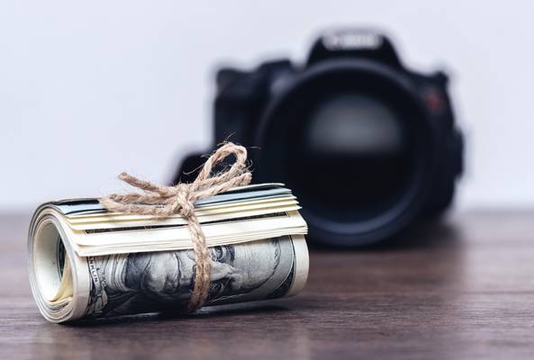 Rolled US dollar bills tied with twine in front of a blurred DSLR camera on wood table