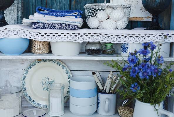 Rustic kitchen shelf with blue floral dishes, utensils, oats, eggs, and wildflowers.