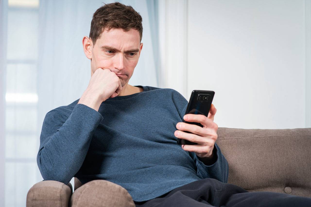 Man in blue shirt sitting on couch looking thoughtfully at smartphone screen