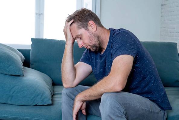Man sitting on blue sofa, looking stressed and overwhelmed, holding head in hand.
