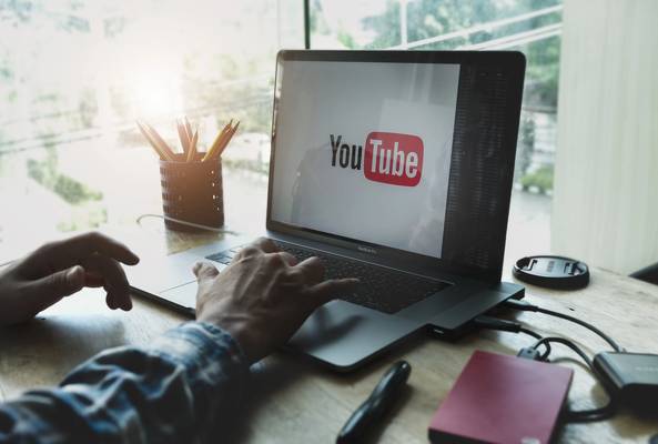Person using MacBook Pro with YouTube logo on screen and pencils on wooden desk.