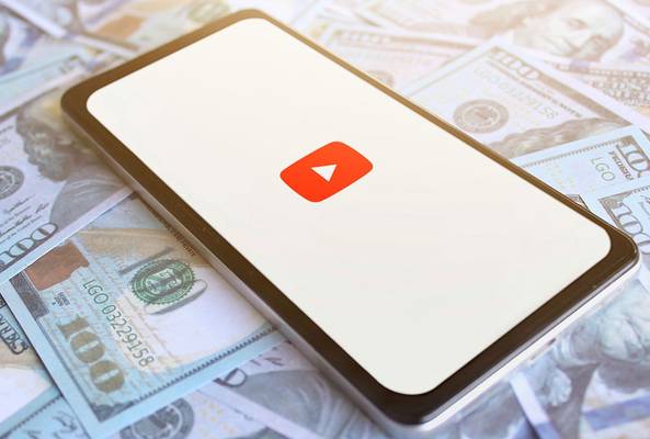 Smartphone showing YouTube logo on screen over scattered hundred dollar bills background.