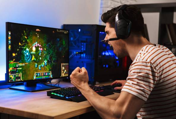 Gamer wearing headset playing intense video game on desktop computer with RGB keyboard.
