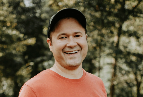 Smiling man wearing a black cap and red shirt outdoors with blurred green background