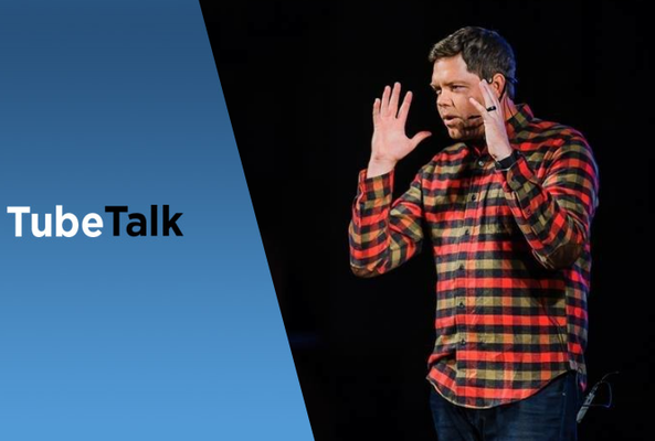 Speaker in red plaid shirt gesturing during TubeTalk presentation on black background
